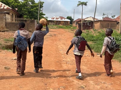 Four young boys, likely students given their uniforms and backpacks, are walking along a dirt road in Barakin Angundi, Nigeria. The boy on the far left wears a plaid shirt and brown pants, carrying a blue backpack with circular patterns. Next to him, a boy in a grey long-sleeved shirt holds a worn, brown soccer ball aloft in his right hand. The third boy from the left, facing away from the camera, wears a plaid shirt with a maroon backpack. The boy on the far right, also facing away, is dressed in a plaid shirt and brown pants, carrying a black backpack. The road is made of reddish-brown dirt with scattered debris. In the background, simple houses with corrugated metal roofs and mud walls are visible, surrounded by green vegetation. The sky is partly cloudy. The scene appears to depict children walking home or to school in a rural Nigerian setting.
