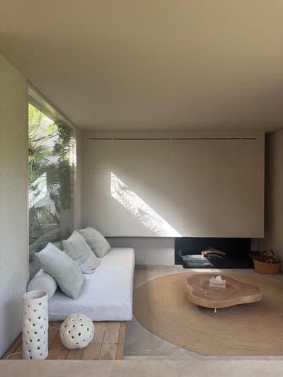 This media file presents a high-angle, top-down view of a modern, minimalist living space with an outdoor, natural element. The scene is bathed in natural light, with strong shadows cast by foliage from the adjacent courtyard, suggesting it is daytime, possibly late morning or early afternoon, given the angle and intensity of the sunlight.

The main subjects are an assortment of objects arranged in a living area. A large, textured, circular rug forms the base of the arrangement. On the rug sits a striking, irregularly shaped wooden coffee table with a live edge, featuring a small stack of white tissues. To the right of the coffee table, a white linen sofa is adorned with several light grey linen cushions. In the foreground, to the right of the sofa, are two unique decorative objects: one is a spherical light fixture with numerous small holes, and the other is a cylindrical fixture with a similar perforated design. These rest on a textured, light-colored floor, possibly terrazzo, adjacent to a wooden surface.

Behind the sofa and coffee table is a dark, recessed area, possibly a fireplace, with glimpses of natural materials like wood or bark in woven baskets above it. The wall behind the sofa is a smooth, neutral-toned surface, marked by a rectangular vent and a subtle square inset. A large window or glass wall to the left reveals a lush courtyard filled with palm trees and other greenery, creating a strong connection between the indoor and outdoor environments. The contrast between the clean lines and muted colors of the interior and the vibrant greenery outside is a notable detail.

The overall impression is one of serenity, comfort, and a harmonious blend of natural and modern elements. The scene is devoid of people, focusing solely on the curated arrangement of objects and the interplay of light and shadow. The location context of Artà, Spain, suggests a warm climate, which aligns with the outdoor greenery visible. There is no visible text in the media file.