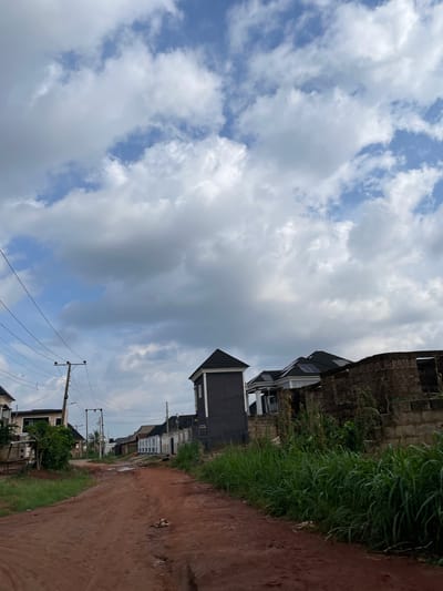 The image depicts a red dirt road in the city of Edopa, Nigeria, leading through a residential area. The sky is filled with scattered white and gray clouds. On either side of the road, there are houses in various stages of construction or completion. Some houses are modern with dark pitched roofs, while others are unfinished, with exposed brick walls. There are utility poles and power lines visible along the road. Lush green vegetation, including tall grass and bushes, grows along the sides of the road and around the properties. The road appears unpaved and somewhat uneven, with puddles of water visible in some sections. The overall impression is of a developing neighborhood.