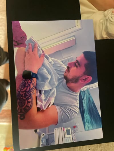 The scene depicts an adult male in an indoor setting, identified as the city of Senden, Germany. The man is seen in profile, facing right, with dark hair and a beard. He wears a light-colored short-sleeved t-shirt. His left arm, adorned with a tattoo featuring roses and a pocket watch, holds a subject wrapped in a light blue towel. A small hand is visible resting on the towel, suggesting the presence of an infant or baby. On his left wrist, he wears a dark smartwatch with a blue band. The background reveals a room with light-colored walls. Visible objects include a white wall-mounted unit, a medical-type monitor device, and a clear dispenser on a shelf in the lower right. A light blue and turquoise artwork depicting waves is mounted on the wall. A window or light source emitting a pinkish hue is visible at the top, and a purple towel or fabric hangs in the upper left background.