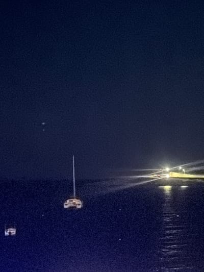 The image shows a nighttime view of Saint Paul's Bay, Malta. On the right side, light reflects off the water's surface. At the top right, a structure appears illuminated with several bright lights, their reflections stretching vertically downward in the water. Slightly below the center of the image, a small object is visible, appearing to be a boat or a small structure on the water, also reflecting light. The left side of the image is primarily a dark, solid color, suggesting the absence of illuminated objects or landmarks in that direction. A few faint dots are visible, likely stars or distant lights.