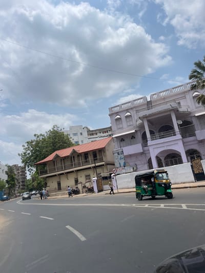 This is a street-level, eye-level, medium shot captured on a sunny day in Ahmedabad, India. The scene depicts a busy street with a light traffic of vehicles, including cars and an auto-rickshaw. On the right side of the street, there is a light purple, multi-story building with ornate white railings and arched windows. Next to it, on the left, is a beige, two-story building with a red tiled roof and wooden balconies. Several people are visible walking on the sidewalk in front of the buildings. The street is paved and marked with white lane lines. Tall trees are present behind the beige building, and in the background, modern high-rise buildings are visible. The sky is partially covered with white, fluffy clouds.