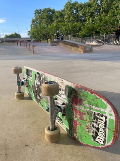 The image captures a well-worn skateboard resting on a concrete surface at a skatepark in Santa Perpètua de Mogoda, Spain, under bright daylight. The skateboard is oriented diagonally across the frame. Its deck is predominantly green and white with significant red and black scuff marks, indicating heavy use.

Prominently displayed on the tail of the skateboard deck is a black sticker with the white text "THRASHER". Other faint text and graphics are visible on the deck, including partially visible words like "AIR M[...]" and some stylized Japanese characters. The skateboard features silver trucks and off-white wheels that show signs of dirt and wear.

In the blurred background, large green trees are visible against a clear blue sky, suggesting good weather conditions. Elements of the skatepark environment are discernible, including concrete ramps, a metal fence, and a metal rail structure. A concrete box or ledge with some graffiti is also present. A blurry figure of a person, possibly a skateboarder, can be seen near the fence in the distance, confirming the active skatepark setting. The strong sunlight casts distinct shadows, indicating a mid-morning or afternoon time of day.