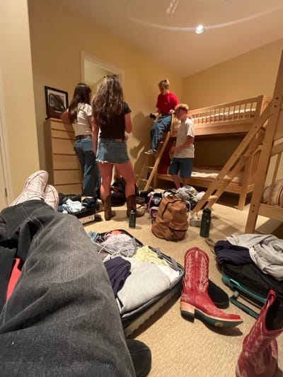 The image shows a living room in Palm Desert, California. Four people are present: two women, one man, and one boy. The two women are standing, one wearing jeans and a white shirt, the other wearing denim shorts and a black shirt. The man is sitting on a bunk bed wearing a red shirt and jeans. The boy is standing on the bottom bunk bed wearing a blue shirt and shorts. Several suitcases, a backpack, clothes, and shoes are scattered around the room, suggesting that the occupants are preparing to travel. The room is decorated with wooden furniture and a bunk bed, with light-colored walls and a large window that lets in natural light. The overall ambiance of the room is casual and relaxed. The image was taken from a low angle, focusing on the suitcases and the person lying on the floor.