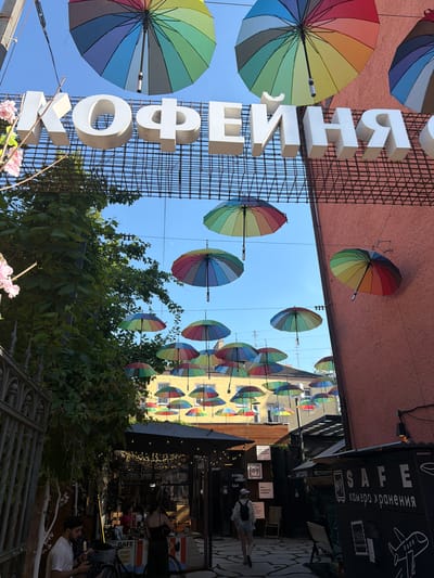 This is a street view in the city of Zelenogradsk, Russia. Above the street, a large sign with white block letters reads "КОФЕЙНЯ" (KOFEYNYA), meaning "coffee shop." Numerous colorful umbrellas are suspended across the street, creating a festive overhead canopy. One large rainbow umbrella is prominently featured at the top of the image. In the foreground, the entrance to a coffee shop is visible, with people seated at tables. The shop's exterior features signs, including one that says "SAFE CAMERA STORAGE." The environment appears to be an outdoor seating area, possibly a cafe or restaurant courtyard, with a paved ground and some greenery. The overall scene suggests a lively and welcoming atmosphere.