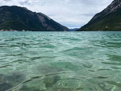 This image captures a low-angle view of the clear, turquoise waters of Achensee in Pertisau, Austria. The foreground is dominated by ripples and small bubbles on the surface of the lake, through which the rocky lakebed is visible. In the mid-ground, the lake stretches towards a distant shoreline. The background features a range of steep, tree-covered mountains that flank the lake. On the left shore, a cluster of buildings, likely part of the village of Pertisau, can be seen nestled at the base of the mountains. The sky above is overcast with diffused light. No people or specific actions are depicted, creating a serene, natural landscape scene.