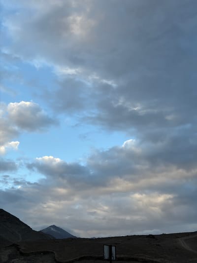 This media file captures a dramatic sky over a mountainous landscape, likely at dusk or dawn, given the soft, diffused lighting and the presence of heavy clouds. The sky is a prominent subject, filled with a mix of dark, brooding storm clouds and lighter, more wispy formations that catch the diminishing or emerging sunlight, creating a sense of depth and movement. A break in the clouds reveals a patch of clear, light blue sky.
To the right of the frame, a steep, dark mountainside dominates the scene. The terrain appears rugged and possibly barren, with visible layers and a rough texture. A winding path is discernible on the mountainside, suggesting human activity or a trail. In the lower right corner, a signpost is partially visible. It is a dark rectangle with some lighter text, but the words are too indistinct to be read clearly.
The overall mood conveyed by the image is one of awe and perhaps a touch of foreboding, due to the imposing nature of the mountains and the turbulent sky. The lack of people and clear man-made structures, other than the sign, places the focus on the raw power of nature. The location context of Chumkar, India, suggests a high-altitude, possibly remote region where such dramatic skies and mountainous terrain are common. The weather appears to be changeable, with the possibility of rain or clearing skies.