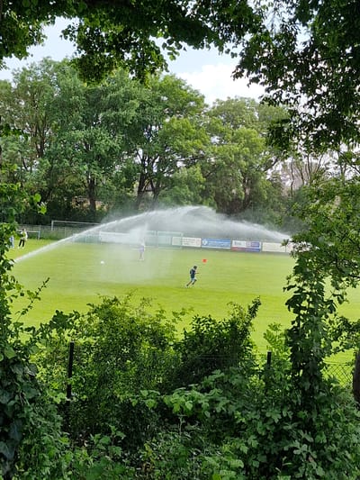 This image shows a soccer field in Bremen, Germany, being watered by sprinklers. The field is green and well-maintained, with a few people on it. There is a soccer goal in the background. The scene is framed by lush green trees.