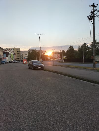 This is a medium-shot, eye-level photograph taken in České Budějovice, Czechia, during sunset. The foreground is dominated by a grey asphalt road. To the left, a street slopes gently upwards, lined with buildings that appear to be shops and apartments. A dark sedan is parked on the side of this street. Further up the street, several other cars are visible.

In the mid-ground, a road curves to the right. The sun is setting behind some trees and buildings in the distance, casting a warm glow across the sky and the scene. The sky is a pale blue with some wispy clouds. Streetlights and utility poles are visible throughout the image, typical of an urban setting.

Notable details include a red triangular yield sign on the right side of the road and a parking sign in front of the buildings on the left. The overall mood of the image is calm and serene, typical of a quiet evening in a city. The time of day is clearly indicated by the low angle of the sun and the warm light. The weather appears clear, with no rain or strong winds suggested.

The visible text includes "REALITNÍ KANCELÁŘ" on a sign on one of the buildings, which translates to "Real Estate Office". Another sign has the word "MOTO" in large letters. The license plate on the parked car reads "BC5 3459".