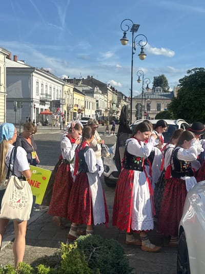 In Nowy Sącz, Poland, a group of young women dressed in traditional folk costumes stand on a street. The costumes feature red skirts with floral patterns, white blouses, and black vests adorned with embroidery. Some of the women have red ribbons in their braided hair. They are standing near parked cars and a white SUV. In the background, there are buildings with ornate architecture, streetlights, and a blue sky with some clouds. One woman on the left is wearing a light blue headscarf and a white t-shirt, carrying a tote bag with various drawings. Another woman is wearing sunglasses and a dark patterned shirt. The scene appears to be a public gathering or event in the city center, possibly a cultural festival or parade.