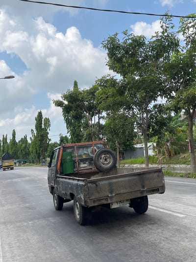 A black pickup truck is driving on a paved road in Kepadangan, East Java. The truck is loaded with a green barrel and a spare tire. The sky is cloudy and blue, with tall trees lining the side of the road. A yellow truck is visible in the distance. The license plate of the pickup truck reads "9744 N". The road has white lines marking the lanes.