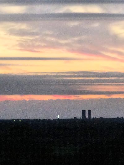 The image depicts a view of the skyline of Bremen, Germany, at sunset. The sky is filled with horizontal bands of clouds in varying shades of gray, pink, and orange, indicating the sun is low on the horizon. In the foreground, a dark, silhouetted landscape stretches across the bottom of the frame, obscuring the ground level details. Two prominent, tall, rectangular structures, likely industrial chimneys or part of a power plant, stand out against the lighter sky. Scattered small lights are visible along the horizon line within the dark silhouette, suggesting urban or industrial lighting. The overall impression is one of a twilight industrial or port area.