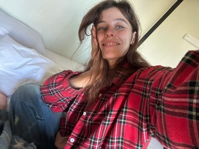 A medium shot shows a young woman with long, brown hair and blue eyes. She is wearing a red plaid shirt and blue jeans. Her right hand is resting on her cheek, and she is smiling. She is sitting in a bed with white pillows. The background is a light-colored wall and a black stripe. The location is not specified.