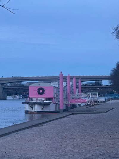A pink and white boat with a large "O" on its side is docked next to a concrete walkway. The boat has several pink cylindrical structures. A bridge spans above the boat, and a body of water is visible. The sky is overcast, suggesting dusk or dawn. Trees are sparsely visible in the background.