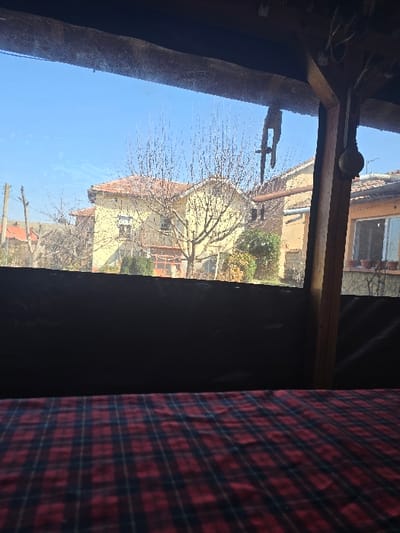 The image depicts an exterior view of a residential street in Petarnitsa, Bulgaria, observed from indoors through a transparent barrier. The foreground shows a red and dark blue plaid tablecloth occupying the bottom portion of the frame. Above the tablecloth, a dark, opaque barrier extends horizontally. The upper part of the interior frame includes a wooden beam at the top and a vertical wooden post on the right. A metallic, hooked object hangs from the top beam towards the right, and a spherical, light-colored object is affixed to the vertical post, with electrical wires visible above it.

Beyond the transparent barrier, a clear, bright blue sky dominates the upper background. Below the sky, several residential buildings are visible. A two-story house with light yellow walls and a terracotta-tiled roof stands centrally, partially obscured by bare deciduous trees. To the right, another building with a similar roofline and lighter walls features a window with several potted plants on its sill. Further to the left, a utility pole with overhead lines is present. Bare deciduous trees are scattered throughout the scene, along with some evergreen foliage near the base of the central yellow house. No individuals or active movements are observed.