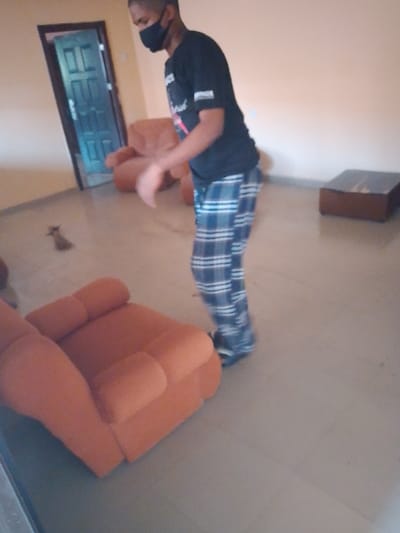 In a living room, a person wearing a black shirt, plaid pants, and a black mask stands near an orange armchair. The room also contains another orange chair, a dark wooden coffee table, and a brown figurine on the floor. A blue door with a dark frame is visible in the background. The room has beige walls and light grey flooring.
