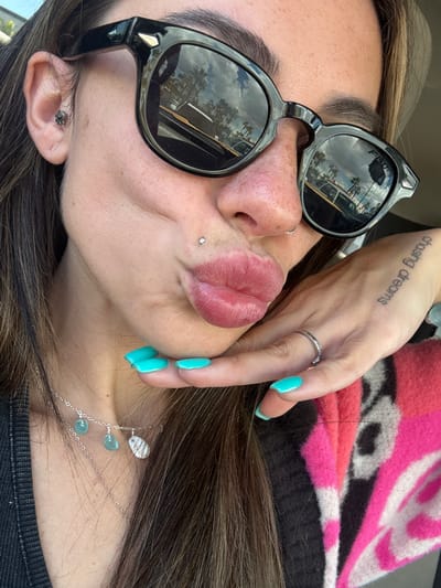 A person, possibly female, is posing in an image. The person is wearing sunglasses that reflect an exterior scene of palm trees, parked cars, and what seems to be buildings under a sunny sky, which is indicative of the city of Long Beach, California, United States. She has pierced ears and a pierced lip. The person is also wearing a ring, and turquoise-colored nail polish on her fingernails. A tattoo that reads "chasing dreams" is visible on the person's right wrist. She is wearing a pink and dark-colored top. There is also a delicate silver necklace around the neck.
