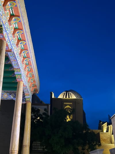 The image depicts the ornate architecture of Samarkand, Uzbekistan, at dusk. The vibrant blue sky dominates the upper portion of the frame, suggesting a clear evening. In the foreground, a brightly colored, intricately decorated wooden structure extends across the lower half of the image. This structure features detailed patterns in blue, yellow, green, and orange, with repeating motifs and what appear to be stylized figures. Wooden beams, possibly part of a balcony or roof, are visible in the lower right.
In the middle ground and background, the warm glow of artificial lights illuminates elements of a building, highlighting its brickwork and a large, domed structure with patterned detailing. A dark green tree stands to the right of this building, its foliage silhouetted against the illuminated architecture and the twilight sky. The overall scene evokes a sense of historical grandeur and cultural richness, likely a tourist site or religious building in Samarkand. There is no visible text in the image.