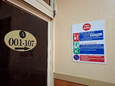 The image shows a fire action sign posted on a wall in Karagita, Kenya (latitude: -0.754, longitude: 36.438). The sign is a blue and red rectangular sign with white lettering. The sign includes a list of actions to take in the event of a fire, including calling the fire safety manager, contacting the fire brigade, and evacuating the building. There is also a sign with the number 001-107, and a black arrow pointing right, on the wall next to the fire action sign. The signs are mounted on a beige-colored wall. The wall is next to a glass door. The room is likely in a commercial or public building.