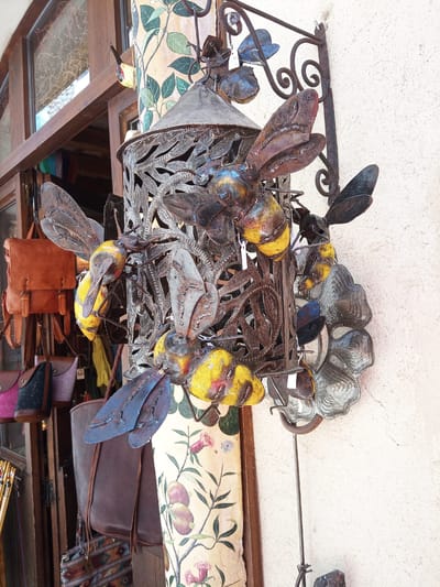 This is a medium shot of a decorative metal sculpture of bees attached to a building's exterior in Besalú, Spain. The sculpture features several metal bees with yellow and black bodies and translucent wings, clustered around a central, ornate metal structure resembling a lantern. The bees are detailed, with visible legs and antennae. Behind the sculpture, a section of a building's facade with a rough, off-white texture is visible. To the left, a wooden window frame with glass panes and a glimpse of a shop interior can be seen, showcasing several leather and fabric bags hanging. A cylindrical decorative element with a floral pattern, featuring green leaves and pink flowers, is positioned to the right of the sculpture. The lighting suggests an outdoor, daytime setting. There are no people visible in the image.