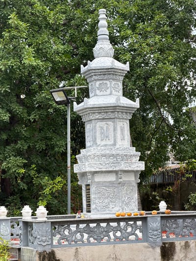 The image displays an ornate, multi-tiered stone stupa or memorial column, carved from light grey stone, set against a backdrop of lush green trees. The structure is intricately detailed with traditional East Asian motifs, including prominent lotus flower carvings. Visible Chinese characters on one central panel read "往生蓮位" (Lotus Seat for Rebirth) and "仙師仙姑" (Immortal Master, Immortal Maiden). Below these, a darker plaque contains further dedicatory text, partially legible as "佛光普照" (Buddha's light shines universally) and "先考" (deceased father), suggesting a memorial purpose.
Dense green foliage from numerous trees fills the background and much of the upper frame, indicating an outdoor, natural setting. A modern LED floodlight is mounted on a metal pole next to the stone structure. To the right, a decorative grey stone railing or gate features elaborate scrollwork and floral patterns, echoing the carvings on the central structure. A string of small, round, yellowish-orange fruits, possibly kumquats, hangs from this railing. In the far right, a textured, light-colored wall is partially visible.
The scene takes place during the daytime, under soft, diffused light, with no people present. The architectural style, carvings, and visible text point to a cultural or religious site in Cam Ranh, Vietnam, conveying an atmosphere of tranquility and cultural heritage.