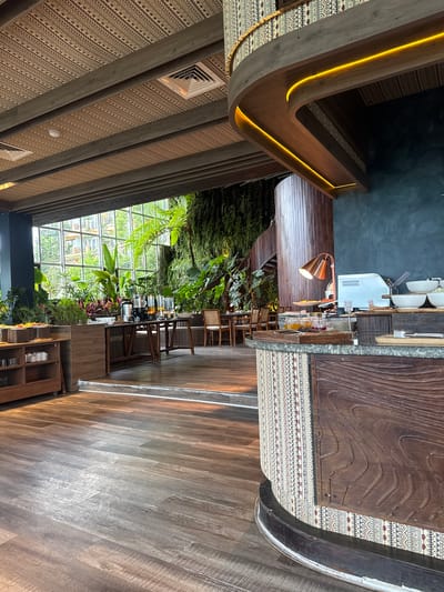 The image shows the interior of a dining establishment in Sa Pa Ward, Vietnam. The architecture features a mix of modern and rustic elements, with exposed wooden beams and patterned ceiling panels. A wall of windows allows natural light to illuminate a lush green space with various plants. A long table is set with glasses and juice dispensers, indicating a buffet or self-serve arrangement. Chairs are arranged along the table. A counter area with a cash register and other equipment is also visible. The floor is made of wood, and overall the scene conveys a natural, upscale dining experience.