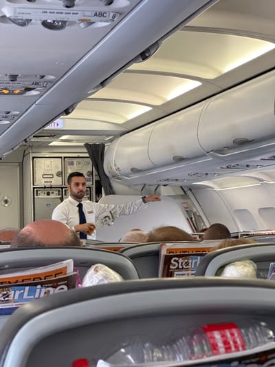 The image depicts the interior of an airplane cabin, likely in flight. In the foreground, rows of passenger seats are visible, with magazines and personal items placed in seat pockets. Several passengers' heads and shoulders are visible, partially obscuring the view of the cabin. In the mid-ground, a flight attendant, dressed in a white shirt and dark tie, is standing and appears to be giving instructions or a safety demonstration, holding an item in his right hand. Behind him, various aircraft equipment and compartments are visible, including storage units labeled "ISRAIR." Overhead, the airplane's ceiling is equipped with lighting and air vents. The overall scene suggests a standard commercial flight environment. While the image is taken inside an airplane, the city of Yehud-Monosson, Israel, is not directly depicted or indicated by any visible landmarks or context within the photograph.