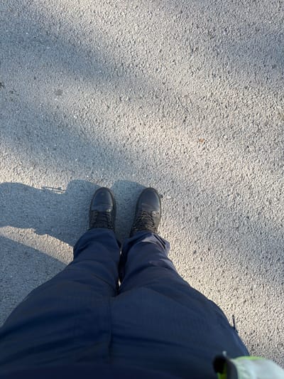 The image, taken in Han Krum, Bulgaria, provides an overhead view of a person's lower body, specifically their legs and feet, on an outdoor asphalt or paved surface. The individual is wearing dark blue pants and black, lace-up boots. A small, light-colored, possibly neon-yellow or green, detail of an item or garment is visible on the far right edge of the frame, near the upper part of the pants.

The ground surface is a textured, light grey asphalt with visible small aggregate stones. Distinct shadows are cast on the ground by the person and possibly other unseen objects, indicating bright sunlight and suggesting it is daytime, likely morning or late afternoon based on the shadow angles. The person appears to be standing or walking on this outdoor surface. There are no other discernible activities, emotions, or visible text in the image.