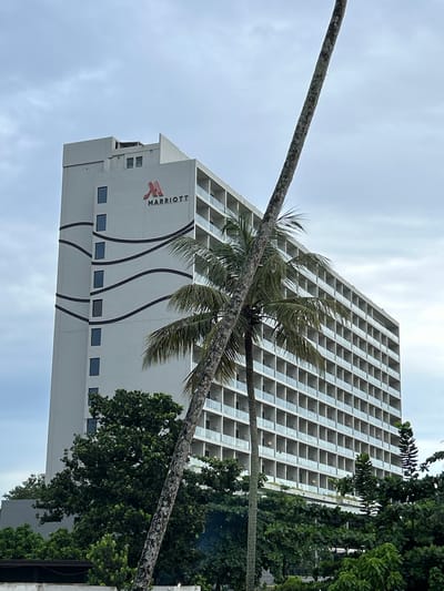 The image shows a tall, multi-story hotel building with the Marriott logo visible on its facade. The building appears to be a modern hotel with many balconies or rooms, each with glass railings. The hotel is situated in a tropical environment, as evidenced by the presence of palm trees and lush green foliage in the foreground and surrounding the building. The sky is overcast, suggesting a cloudy day. The image is taken from a low angle, looking up at the building and the palm tree trunk that dominates the right side of the frame. The location is specified as Peleana, Sri Lanka.
