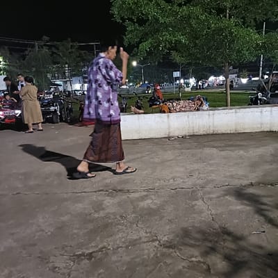 The image shows a street scene in Kaliwungu, Indonesia (latitude: -6.962, longitude: 110.253). A woman in a patterned dress walks along a concrete sidewalk in the foreground. The sidewalk runs parallel to a low concrete wall. A group of people stand in the background. Some are talking, some are looking at something off to the side. Several motorcycles are parked near the curb. A woman lies on the sidewalk in the distance, possibly sleeping. The sky is dark. The scene is illuminated by streetlights.