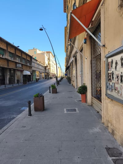 The image depicts a street scene in Livorno, Italy, specifically on Via Grande, characterized by its architecture and paving stones. The perspective is from the pedestrian walkway, looking down the street, which is lined with buildings on both sides. The buildings feature commercial establishments on the ground floor, with shops displaying their wares, some with awnings. The street itself is paved with asphalt and marked with traffic lanes. The pedestrian walkway is paved with grey flagstones and is punctuated by black bollards and planters containing greenery. A tall, slender streetlamp curves over the walkway. The sky is a clear, bright blue, suggesting a sunny day. The overall impression is of a quiet urban environment, possibly in the morning or late afternoon given the gentle shadows.