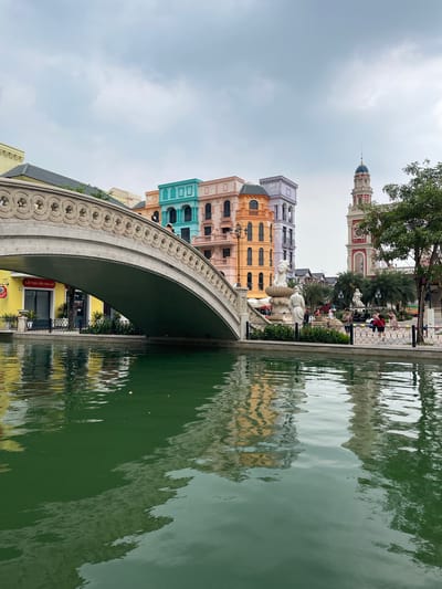 The image depicts a picturesque canal scene in the city of Nghĩa Trụ Commune, Vietnam. A stone bridge with ornate carvings spans the calm, green water, reflecting the colorful buildings on the opposite bank. These buildings feature a distinct architectural style with pastel-colored facades in shades of yellow, orange, teal, and lavender. To the right of the canal, a tall, red and white tower with a blue-domed top rises against a cloudy sky. In the middle ground, a fountain with statues is visible, and several people are strolling along the paved walkway beside the canal. Palm trees and lush greenery add to the tropical ambiance of the location.