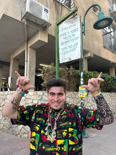A young adult male with dark, voluminous hair and prominent reddish-brown eyebrows smiles directly at the camera. He wears a short-sleeved black shirt patterned with green, yellow, and red squares featuring images of Bob Marley and "ROOTS REGGAE" text. Multiple gold-colored chains with various pendants and colorful tassels adorn his neck. On his left arm, he wears several beaded bracelets with colorful pom-poms and a sleeve or temporary tattoo depicting a marijuana leaf within a peace symbol. His right arm displays a black and white tribal-patterned sleeve and multiple wristbands, including yellow and Rasta-colored ones. He holds both hands up, forming modified "shaka" gestures with thumbs pointed inwards and index fingers extended upwards.
The scene is an outdoor setting in Bnei Brak, Israel, during daylight. Behind the man, a light brown multi-story building with a rough texture is visible, featuring an air conditioning unit and a drainage pipe on its facade, along with barred windows. To his left (viewer's right), a green metal pole supports a white rectangular sign with Hebrew text. The sign reads, "בס"ד" (With God's help), and the main message states: "Respected guests, in this neighborhood, all boundaries of modesty and holiness are maintained. Please, respect us by dressing modestly. Neighborhood Committee." Above the sign, a green street lamp with a downward-facing fixture extends from the pole. A low stone wall with irregular light-colored stones runs behind the man, and green vegetation is visible beyond it. The ground is a paved sidewalk composed of reddish-brown and gray interlocking pavers.