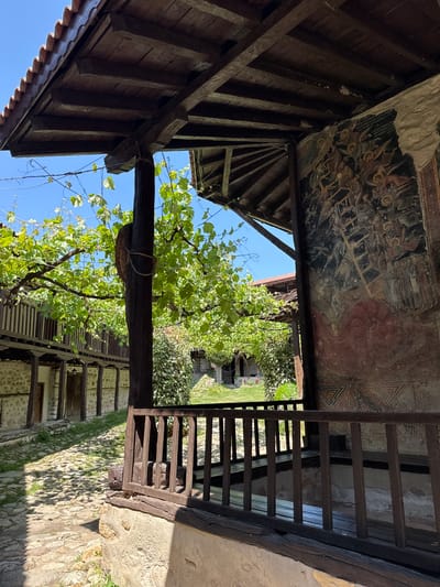The image presents a detailed view of a historical architectural complex in Sandanski, Bulgaria, observed from a shaded, elevated wooden platform. In the immediate foreground on the right, a dark wooden railing supported by a stone base defines the edge of the platform. Behind this railing, a large, weathered fresco, featuring numerous figures with halos and a prominent ladder-like element, covers a substantial portion of a stuccoed wall. The fresco exhibits significant pigment loss and degradation, revealing underlying plaster and stone, and displays muted earth tones. Below the main fresco, a geometric pattern is discernible.
A thick, dark wooden support post rises vertically, heavily entwined with a vibrant green grapevine that forms a natural canopy over a portion of the courtyard. Overhead, a dark wooden ceiling structure with exposed beams and planks is visible, with terracotta roof tiles partially seen on the upper left. To the left of the central wooden post, a sun-dappled cobblestone courtyard stretches into the background, flanked by a stone and timber building. This building features a covered walkway on its ground floor, supported by wooden posts, leading to several dark wooden doors. A wooden railing marks the second story of this structure. Further into the background, lush green foliage, trees, and glimpses of another building with a red-tiled roof are visible beneath a clear blue sky. No people are present in the scene.