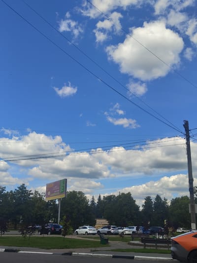 This media file depicts a scene on a street in Ramenskoye, Russia. The image is taken from a low angle, looking up at a bright blue sky filled with fluffy white clouds. A prominent wooden utility pole with numerous wires stretches across the frame, angled diagonally from the bottom left to the top right. These wires create a visual pattern against the sky.

On the right side of the frame, a sidewalk and a road are visible, bordered by a strip of green grass and trees. Several cars are parked along the side of the road, including a white car, a blue SUV, and other vehicles. One car, an orange and gray vehicle, is partially visible in the top right corner. A bus stop bench is also present.

A notable detail is a large advertising billboard, supported by a pole, situated on the right side of the image. The billboard has text in Russian, featuring words like "SANTEL", "АМЕРИКАНС", and a phone number. The time of day appears to be daytime, given the bright sunlight and clear sky, possibly late morning or early afternoon. The weather seems pleasant, with a mix of sun and clouds. The overall impression is a typical street scene on a clear, cloudy day in a Russian town.