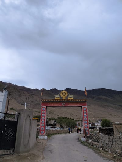 The image shows an arched gateway in Khardung, India, leading to a village. The sky is overcast with gray clouds. The gateway is ornate, with red and gold detailing, and features Buddhist iconography, including a Dharma wheel and a deer. There are also flags flying from the top of the gateway. The road leading through the gateway is paved and appears to be a village road. Several people are visible walking in the distance, and a few buildings are present on either side of the road. The surrounding landscape consists of barren, rocky hills. The overall impression is one of a remote, mountainous region with a strong Buddhist cultural presence.