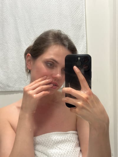 A woman stands in what appears to be a bathroom, evident by the tiled wall and white horizontal molding along the upper part of the wall. She is holding a black smartphone up to take a selfie. The camera is covering part of her face. Her skin appears wet, and a white towel is wrapped around her torso. Her hair is pulled back. The location, Kranj, Slovenia, cannot be confirmed based on this image.