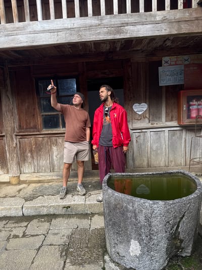 Two men stand near a stone trough filled with water in Sà Phìn Commune, Vietnam. The man on the left points towards a wooden wall with a window that has graffiti on it. Various signs, including "No Smoking" and fire safety instructions, are mounted on the wall. The other man stands beside the trough, wearing a red jacket and sunglasses. The ground is paved with stone. A wooden staircase is visible to the left.
