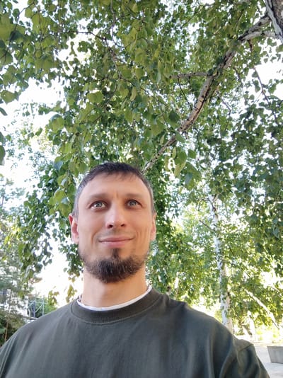 A man is looking up and to his right, with a slight smile on his face. He has short, dark hair with some graying at the temples and a goatee. He is wearing a dark green crewneck sweatshirt with a white t-shirt collar visible underneath. The man is outdoors, with a backdrop of lush green leaves and tree trunks, suggesting a natural or park-like setting. The light filtering through the leaves indicates it is daytime, likely late morning or afternoon, with sunny weather. The presence of birch trees with their distinctive white bark is a notable detail. The overall impression is one of a relaxed and pleasant outdoor moment. No visible text is present in the image. This setting is consistent with Petropavl, Kazakhstan.