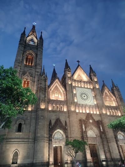 The image shows the Expiatory Temple (Templo Expiatorio del Santísimo Sacramento) in Guadalajara, Mexico, at dusk. The Gothic-style church is illuminated, highlighting its architectural details, including spires, crosses, rose window, and arched doorways. The sky is a deep blue with scattered clouds. Green trees are visible on either side of the building. At the base of the church, on the right side of the frame, a few people are partially visible.