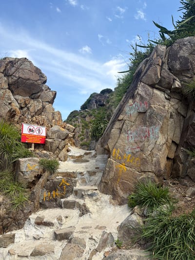 The image depicts a rocky pathway with stone steps leading upwards between large, weathered rock formations. The path is partially paved with concrete and irregular stones, with patches of yellow paint or markers on some rocks and steps. Green grass and vegetation grow sparsely around the rocks.
On the left side of the path, a red and white sign is posted on a rock. The sign features symbols and text in multiple languages, including Vietnamese and English. The English text reads, "Please do not bring food and drinks and litter inside the cave." Below this, there is text in Cyrillic script.
The rocks themselves show evidence of graffiti, with red and blue markings on the larger rock face to the right of the path. The sky above is blue with scattered white clouds. In the background, the path continues to ascend into a hilly, vegetated area. The overall scene suggests a natural, somewhat rugged outdoor location likely used as a hiking trail or access route to a natural feature. The location is identified as being in the city of Vĩnh Lương, Vietnam.