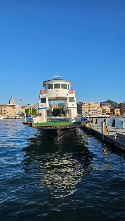 The image captures a vibrant, clear day at a harbor, consistent with a scenic location like Como, Italy. A white ferry, featuring blue accents and a distinctive upper deck with numerous windows, is prominently docked in the foreground. Several orange life preservers are affixed to its sides. The boat's reflection, along with patterns of sunlight, shimmers across the rippling, dark blue water.
Along the distant shore, a cluster of multi-story buildings with various architectural styles and light-colored facades rise towards a gentle hillside. Notable structures include a building with a large dome or cupola and a construction crane. Green hills and mountains form a natural backdrop behind the urban development. The sky above is a brilliant, cloudless blue, indicating excellent weather.
The scene appears quiet, with no immediate human activity visible on the boat or along the waterfront. The ferry seems to be at rest, possibly awaiting passengers or a future departure. No clearly legible text is discernible on the boat, buildings, or other elements within the frame.
