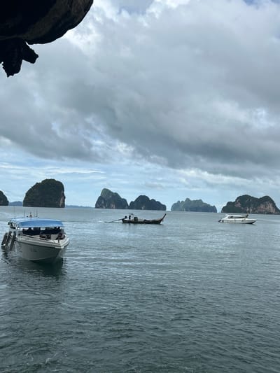 In Tambon Krasom, Thailand, a wide shot captures a scenic view of the sea under a cloudy sky. In the foreground, a white speed boat with a blue canopy is docked. Further out, a longtail boat with two people is visible, and to the right, another white speed boat is anchored. The background features several lush, green limestone karsts rising from the calm sea. The water is a deep blue, with gentle ripples on the surface, and the sky is filled with dramatic grey clouds. A large, dark rock formation hangs from the top left corner of the frame, casting a shadow.