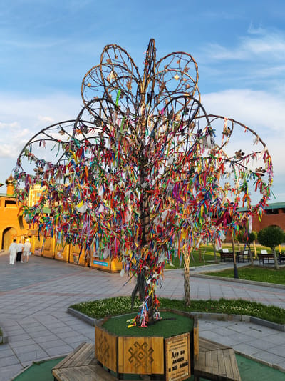 This media file captures a vibrant outdoor scene featuring a decorative tree adorned with numerous colorful ribbons. The tree, made of twisted metal branches, is a prominent central object, its base surrounded by a decorative wooden planter. The planter has an inscription, partially visible, that reads "Tree of love and wish fulfillment" in English, along with Cyrillic text.

In the background, there are several wooden market stalls, suggesting a public space or a fair. Two people, dressed in white, are walking away from the camera on a paved walkway. Further in the distance, there are brick buildings with arched entrances and golden domes, hinting at a historical or cultural site, consistent with the location context of Yoshkar-Ola, Russia. The sky is a clear blue with wispy clouds, indicating it is daytime, likely late afternoon due to the warm lighting. The overall mood is cheerful and festive, amplified by the bright ribbons and the pleasant weather.