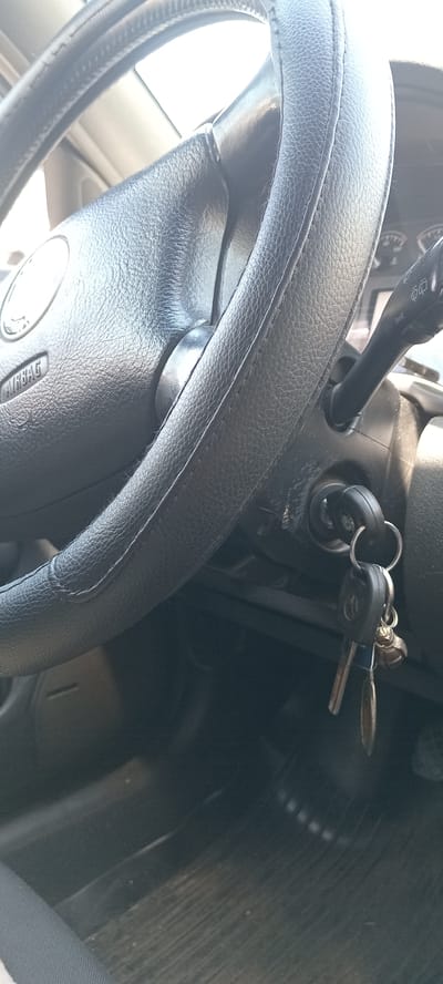 The image displays a close-up view of a car's interior, specifically focusing on the steering wheel and ignition. The steering wheel is black and appears to be covered with a textured material. On the left side of the steering wheel, the word "AIRBAG" is visible. To the right of the steering wheel, an ignition key with a keychain is inserted into the ignition slot. The keychain has a metallic ring and a black fob, which appears to have a car logo on it, possibly Volkswagen based on the visible symbol on one of the keys. A turn signal lever is also partially visible to the right of the steering wheel. The background shows parts of the car's dashboard and the floor mats, which are dark and ribbed. There are no people visible in the image. The lighting suggests it is daytime, with light coming from the right, casting some shadows. The location of Koviahy, Ukraine is not identifiable from the visual information provided in the image.