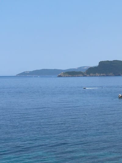 The image depicts a serene coastal scene in Pržno, Montenegro, under a clear blue sky. The foreground is dominated by the calm, blue waters of the Adriatic Sea, reflecting the bright daylight. The water's surface is textured with gentle ripples.
In the mid-ground, a narrow strip of land is visible, characterized by lush green, tree-covered hills that rise steeply from the water's edge. A small town or village is nestled along the coastline, with buildings peeking through the dense foliage. This suggests a populated area, likely a resort or a residential community, integrated with the natural landscape.
Further out at sea, a small, white boat is sailing, leaving a thin wake behind it, indicating movement. A small, dark rock or island is also visible in the water, adding to the natural features of the seascape. The overall atmosphere is peaceful and tranquil, suggesting a warm, clear day, possibly late morning or early afternoon, given the bright lighting. The location cues, such as the mountainous coastline and the Adriatic Sea, firmly place the scene in Pržno, Montenegro. There is no visible text in the image.