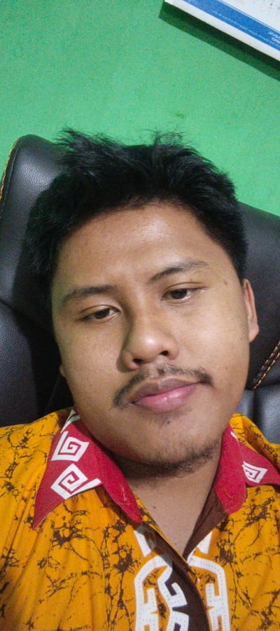 The image is a close-up selfie of a young man, likely in his early 20s, sitting in a black leather chair. He has dark hair, a mustache, and a faint goatee. He is wearing a yellow batik shirt with a red collar that features a white spiral pattern. The background is a solid green wall, and a portion of a desk with papers is visible in the upper right corner. The man appears to be looking directly at the camera with a neutral expression. The lighting is moderate, with some visible grain in the image. There is no specific indication of the location within Sukabumi, Indonesia, other than what might be inferred from the man's appearance and attire, which could be consistent with Indonesian culture. The image captures a simple, personal moment.