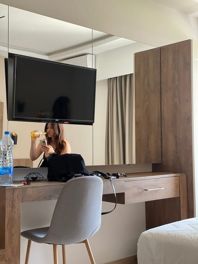 A woman is reflected in a mirror in a room, holding a bottle and a phone. The room appears to be a hotel room in Donji Štoj, Montenegro. A large television is mounted on the wall above a mirrored surface. A wooden dresser or vanity with a drawer is positioned beneath the mirror. A grey upholstered chair with wooden legs sits in front of the dresser. To the left of the dresser, a bottle of water and a black backpack are visible. To the right, a portion of a white bed is in view. The walls are a neutral color, and beige curtains are drawn on the right side of the mirror. An air conditioning unit is mounted on the wall above the television.