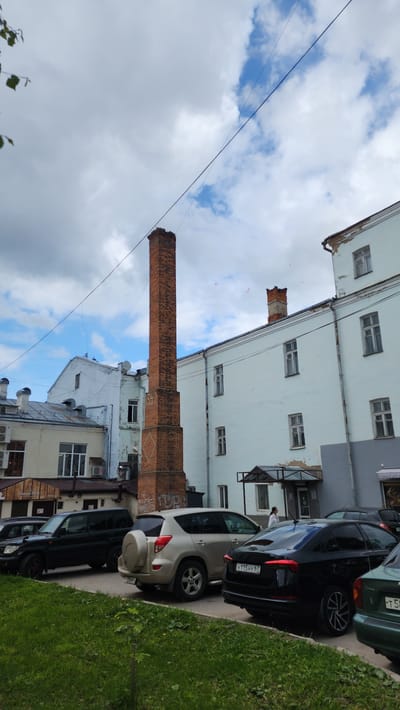 The image depicts a street scene in Smolensk, Russia, featuring a tall brick smokestack rising above a cluster of buildings. The buildings vary in style and color, ranging from white to gray, with multiple windows and architectural details. Cars are parked along the side of the road, including a silver Toyota RAV4 with the license plate "H 115 HY 67". Lush green grass grows along the edge of the street. The sky is partly cloudy with patches of blue. Power lines run diagonally across the sky. The smokestack appears old and weathered, with some damage at the top.