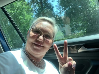 A woman with blonde hair and glasses is taking a selfie inside a car. She is wearing a white t-shirt and a beaded necklace. Her right hand is raised, forming a peace sign. The background shows trees and a building through the car window. The scene appears to be outdoors in a rural or suburban area, possibly Dal'neye Konstantinovo, Russia, given the context of the request.